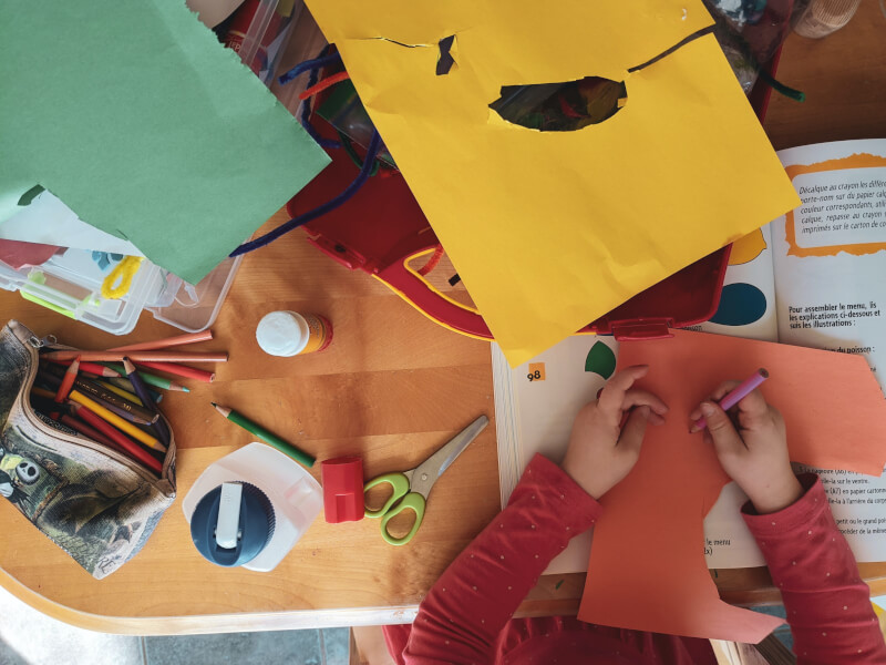 Child drawing on coloured paper surrounded by pens and paper